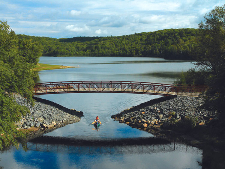 Vue aérienne d'un kayakiste pagayant sous un pont dans un lac, entouré d'arbres