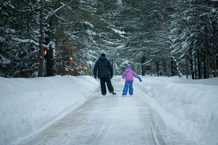 An adult and a child ice skate along a forested skate trail.