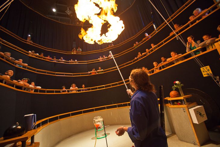 A woman blows out fire as an audience watches the science demonstration. 