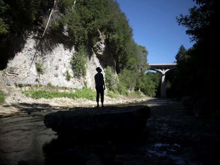 Un homme se tient sur un grand rocher le long d’un cours d’eau et regarde les forêts et les falaises.