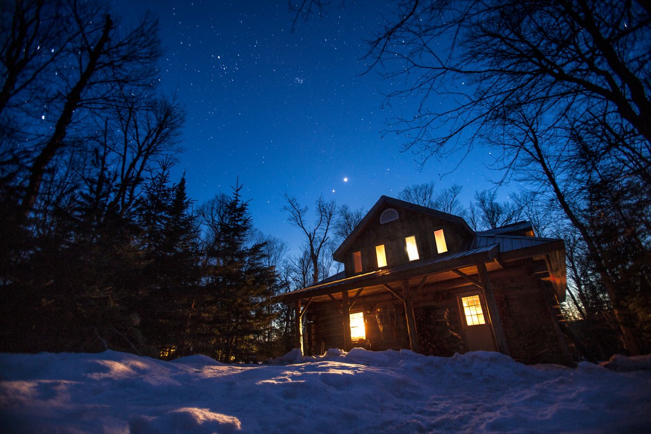 A log home surrounded by snow lit up against the starry evening sky