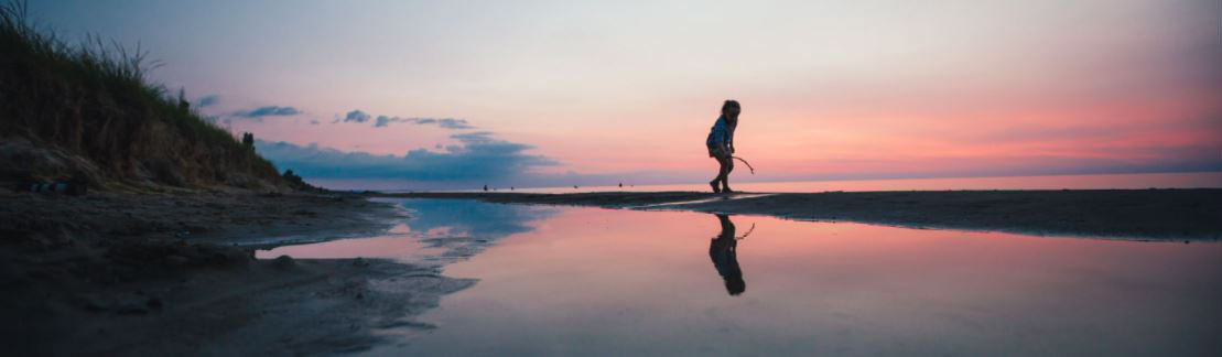 A child playing with a stick on a beach during the pink sunset filled sky.