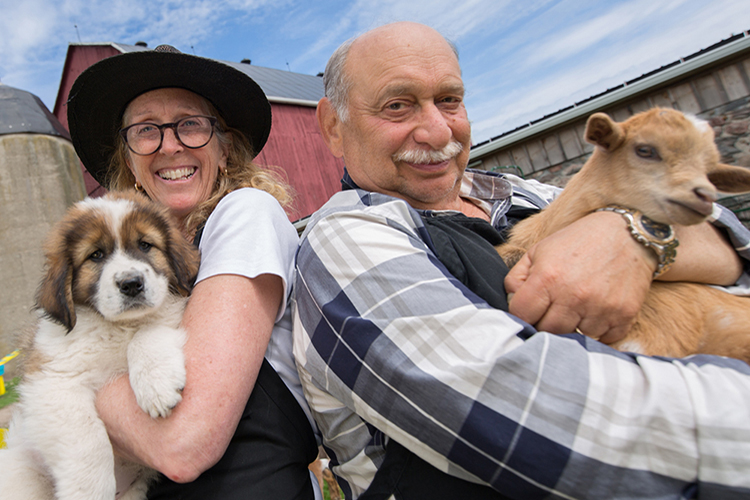 A woman holding a puppy and a man holding a goat stand in front of a barn.