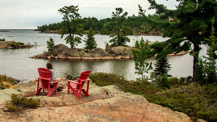 A woman sitting in a red Muskoka chair beside an empty red chair a top a rockface surrounded by water.