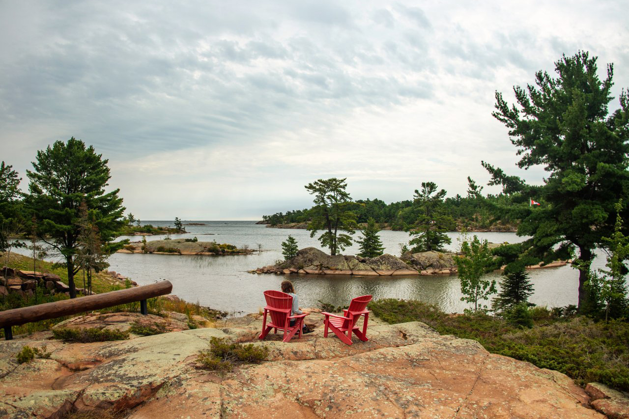 A person sitting in one of two red Muskoka chairs sit on top of a rock looking at a body of water