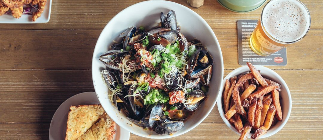 A large round bowl full of muscles, vegetables and shredded cheese, with a side of fries, garlic bread, breaded shrimp and a full glass of amber beer 