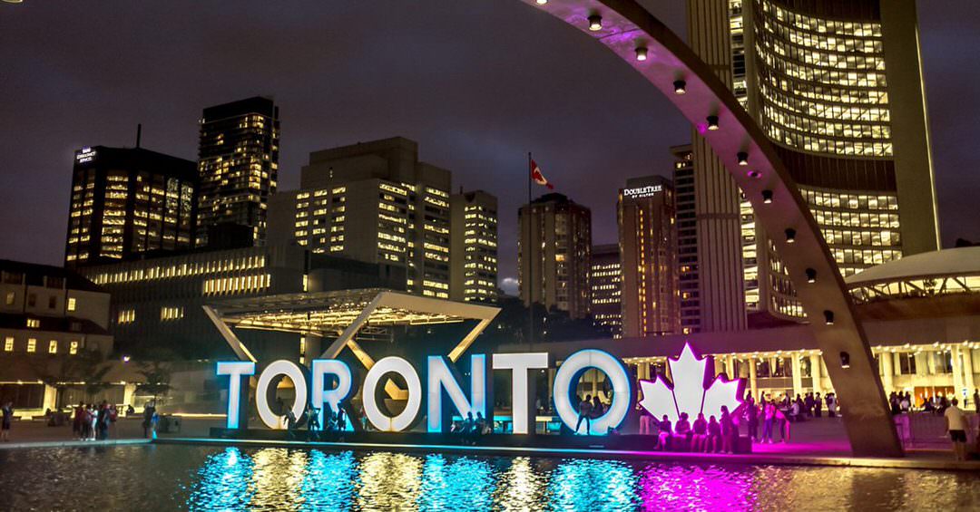 People enjoying the evening in front of and around the large outdoor Toronto sign , which is lit with multicolours as the center piece for the city hall, surrounded with tall buildings.