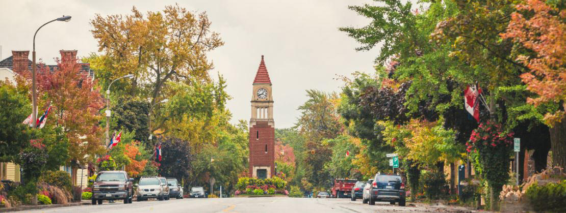 A clock tower divides the two-way road, where on either side are parked cars and houses nestled among trees.