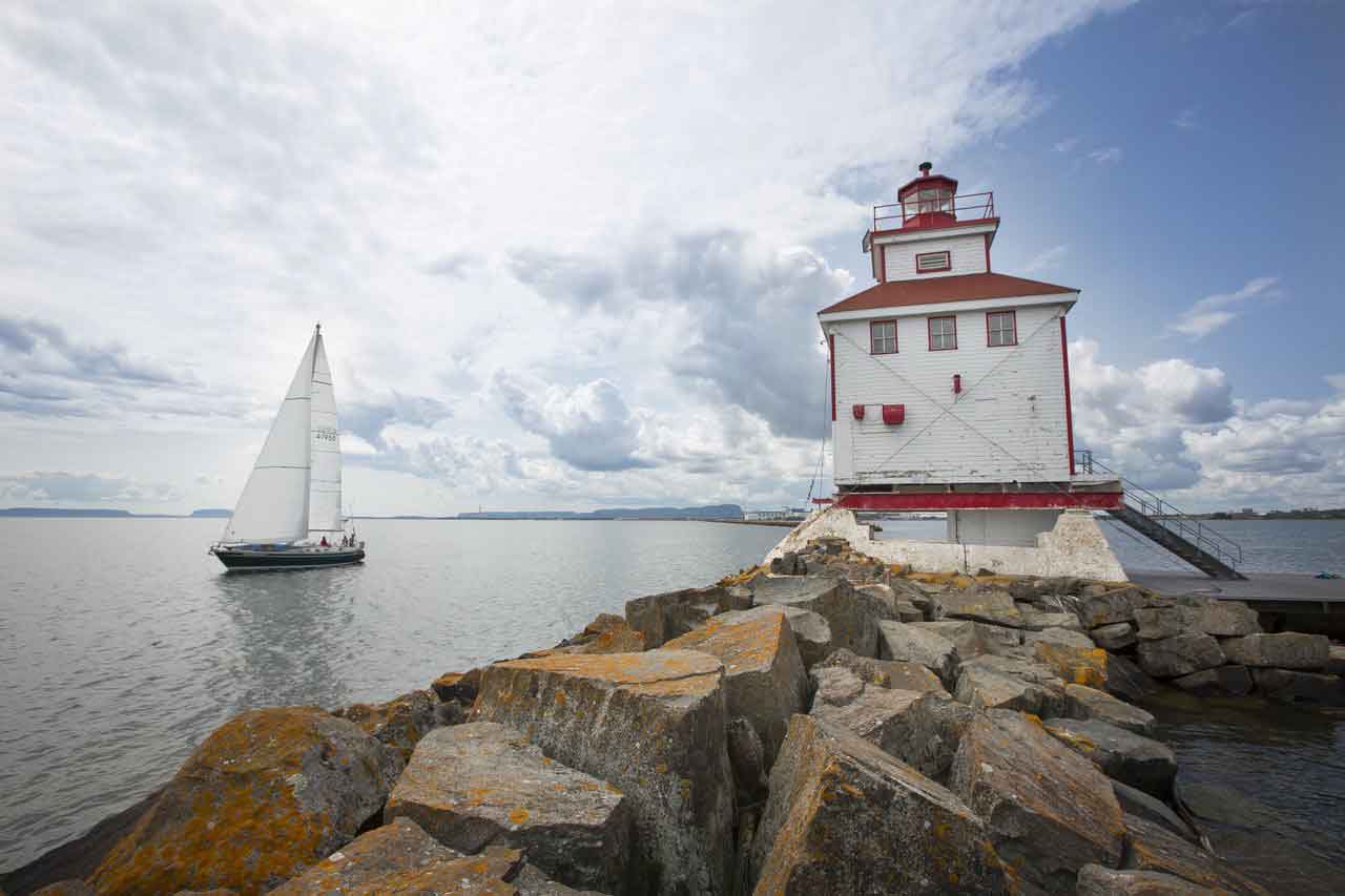 A sail boat sails past a white and red lighthouse atop a rocky coastline