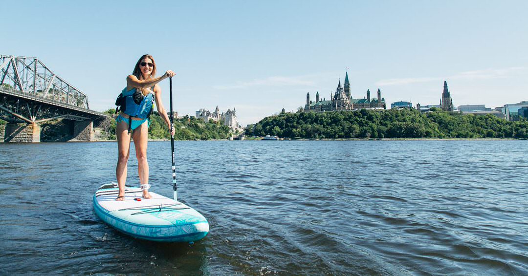 Une femme fait de la planche à pagaie sur l’eau avec derrière elle les édifices du Parlement au haut de la colline.