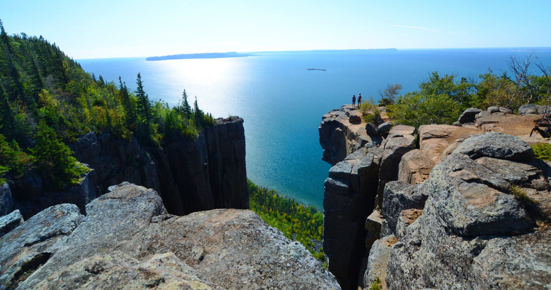 Two people standing in the distance on the edge of jagged cliffs admiring a large body of water and a bright big blue sky