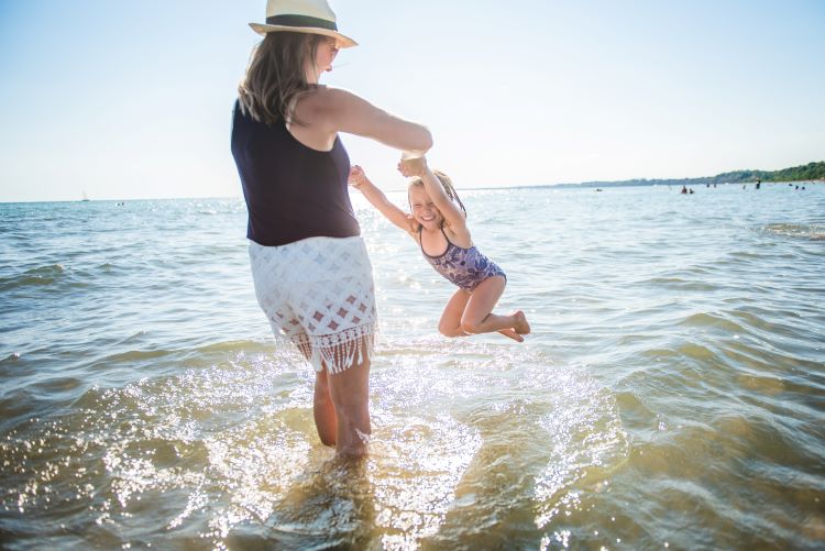 Une mère, les pieds dans l’eau, pivote en tenant les bras de sa fille.