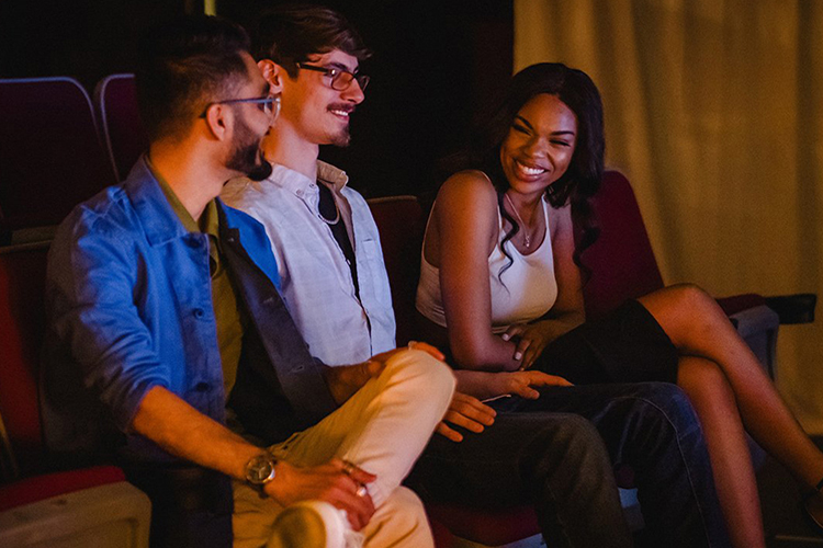 Three people sit in theatre seats awaiting the start of a show.