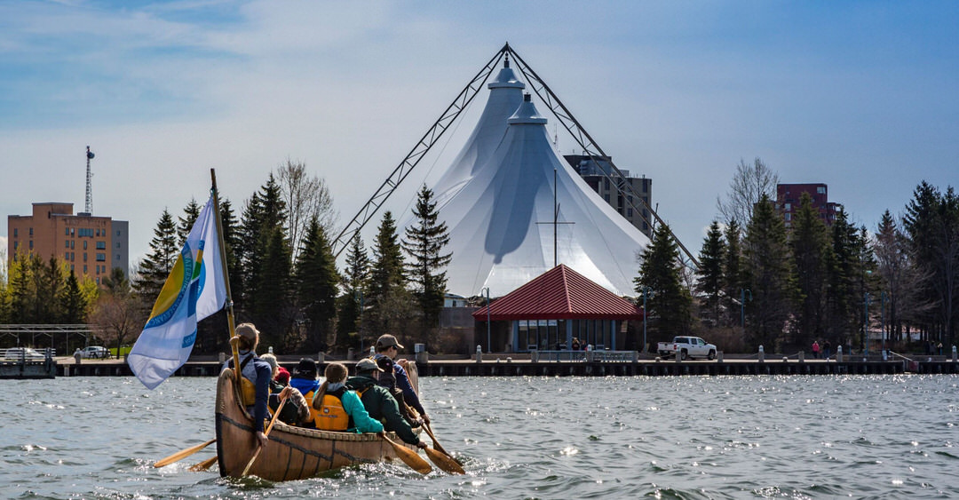 A group of people in a birch bark canoe rowing towards a white tented pavilion located on the waterfront