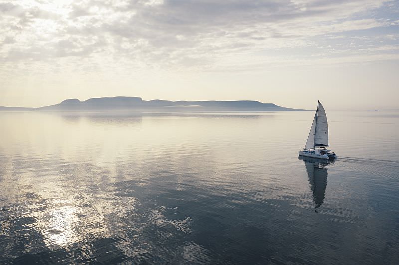 Reflections on water of a catamaran sailing towards hills in the distance as the sky is illuminated a purple gray.