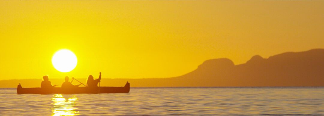 Trois personnes traverse le lac en canot, le géant endormi se découpant sur le coucher du soleil jaune vif en arrière-plan 