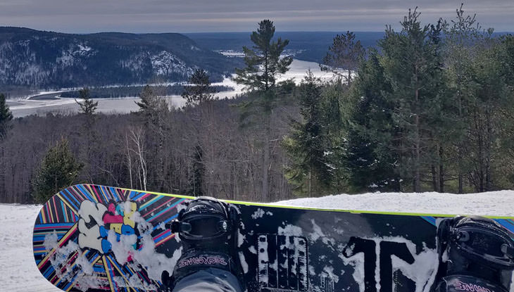 A snowboarder’s view of a forest, lake and mountains from the top of a hill.