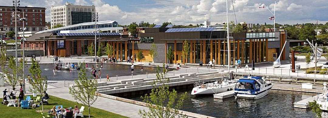 A waterfront full of people lounging, walking and wading in a fountain pool, including an area to dock boats, with access to a community building