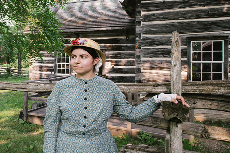 A woman in costume stands in front of a recreated 19th-century home.