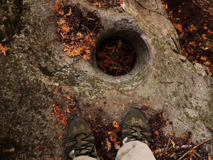 A hiker stands on top of a large rock with a hole in it. 